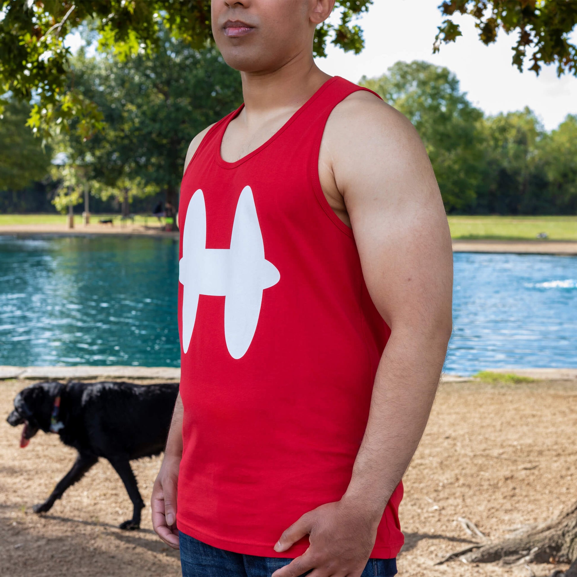 Side profile of man wearing red Houmane tank top with white H logo. Dog park in background.